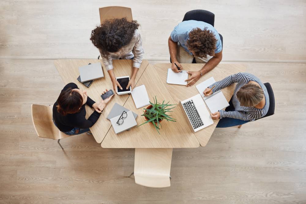 Slika iz ptičije perspektive radnog stola u kancelariji za kojim sede 4 osobe i imaju timski sastanak